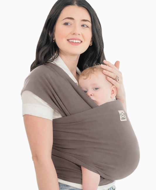 A woman with long dark hair smiles while carrying a baby in the keababies Baby Wrap Carrier - Copper Gray. The baby looks out comfortably as she gently supports their head with her hand.