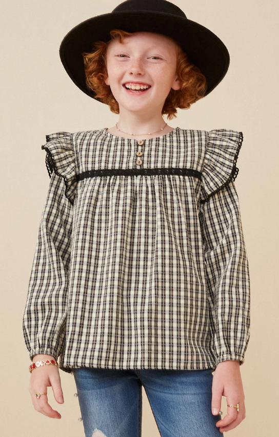 A smiling child with curly red hair wears the Hayden LA Checkered Lace Trim Ruffled Shoulder Top, styled with a wide-brim black hat and blue jeans, standing against a neutral background.