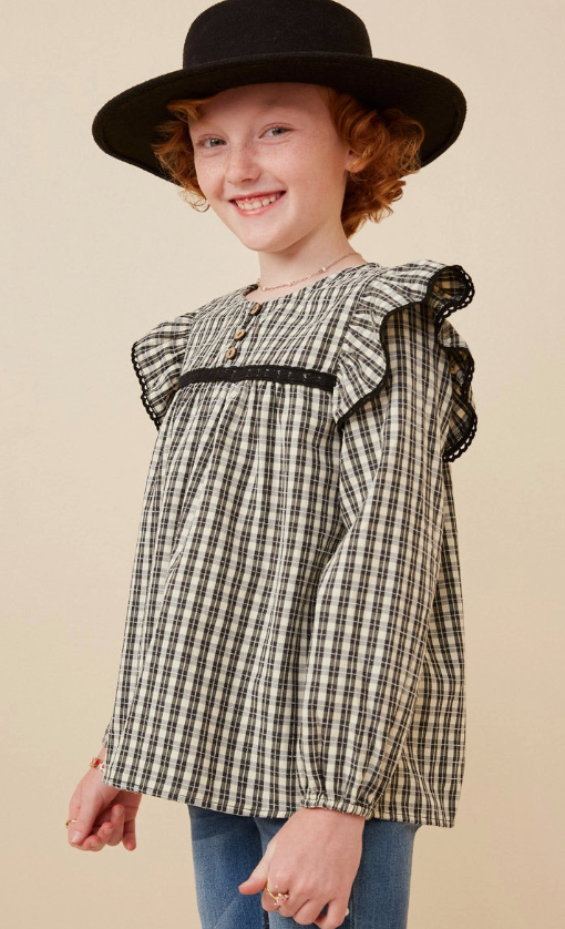 A smiling child with curly red hair wears the Hayden LA Checkered Lace Trim Ruffled Shoulder Top and a black wide-brimmed hat, standing against a neutral background.