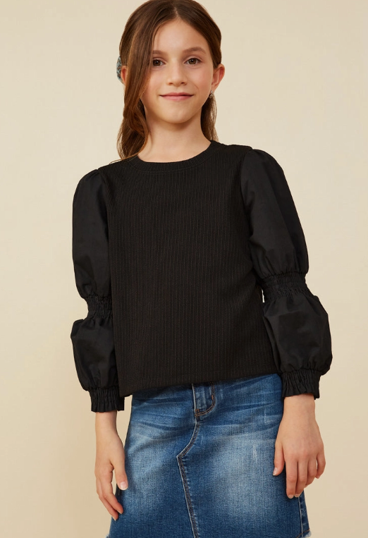 A young girl with long brown hair smiles at the camera, wearing the Hayden LA Puff Sleeve Textured Knit Top in black and a blue denim skirt, standing against a beige background.
