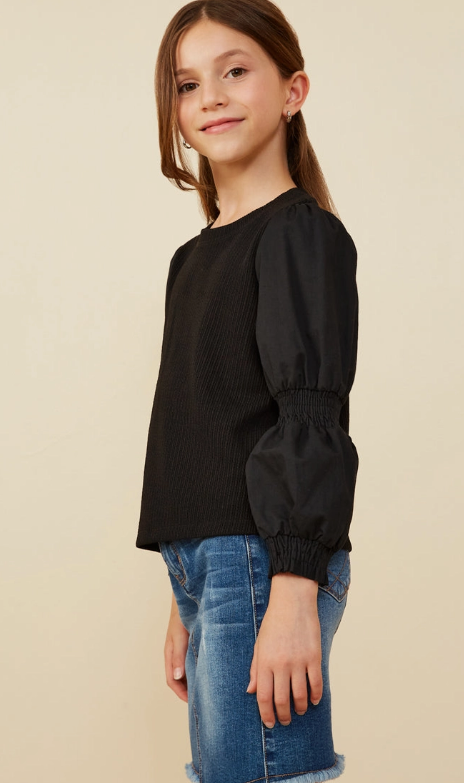 A young girl with long brown hair smiles slightly, wearing the Puff Sleeve Textured Knit Top by Hayden LA and blue denim shorts, standing against a plain light beige background.