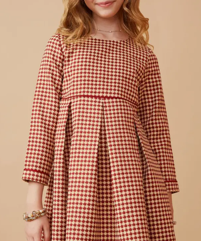 A girl models the Hayden LA Classic Red Houndstooth Pleated Dress with long sleeves and a red-and-cream houndstooth pattern, standing against a beige background. She also wears a bracelet on her left wrist.