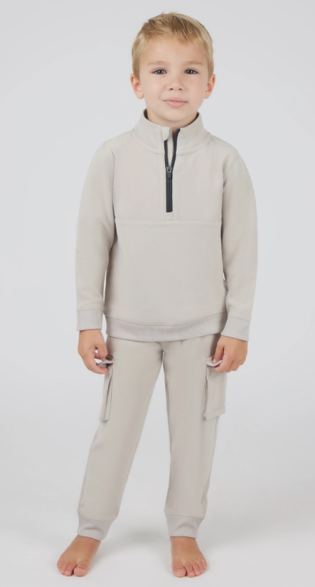 A young boy with short blond hair stands barefoot, smiling slightly in the Cozy Zip-Up Cargo Set by 90 Degree by Reflex & Yogalicious—light beige long-sleeve pullover with black zipper and matching cargo pants—against a plain white background.