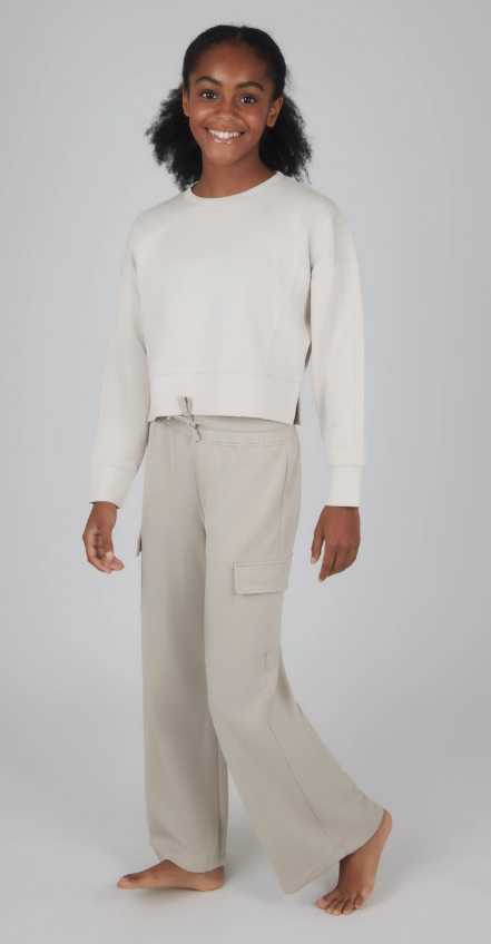 A young girl with dark curly pigtails smiles while wearing the Weekend Comfort Cargo Set from 90 Degree by Reflex & Yogalicious, featuring a light beige long-sleeve sweatshirt and loose cargo pants, standing barefoot against a plain gray background.