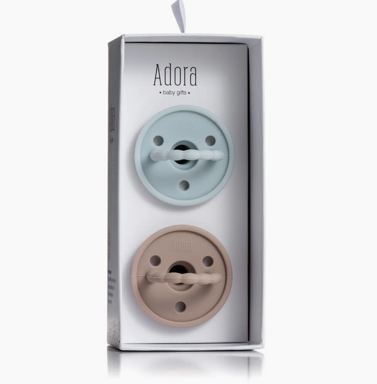The Adora Saddle Adora Pacifier Set - Sky includes two pacifiers, one light blue and one beige, displayed vertically in clear packaging labeled with the Adora logo and "baby gifts" on the front.