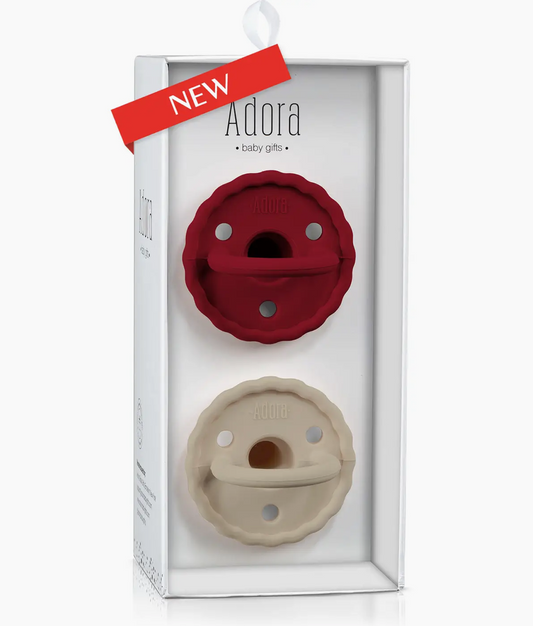 The Adora Saddle Pacifier Set - Scarlet features two silicone pacifiers, one red and one cream, displayed vertically in a box with a "NEW" red label at the top left of the packaging.