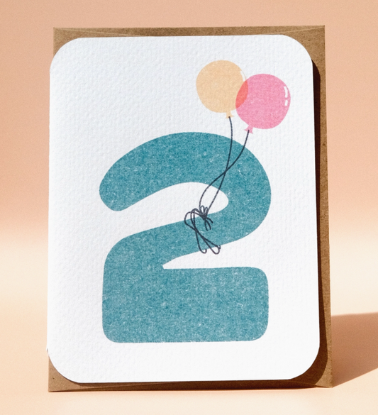 The 2nd Birthday Card by Paper Chain Studios features a large blue number 2, peach and pink balloons attached by black string, rounded corners, and a brown backing.