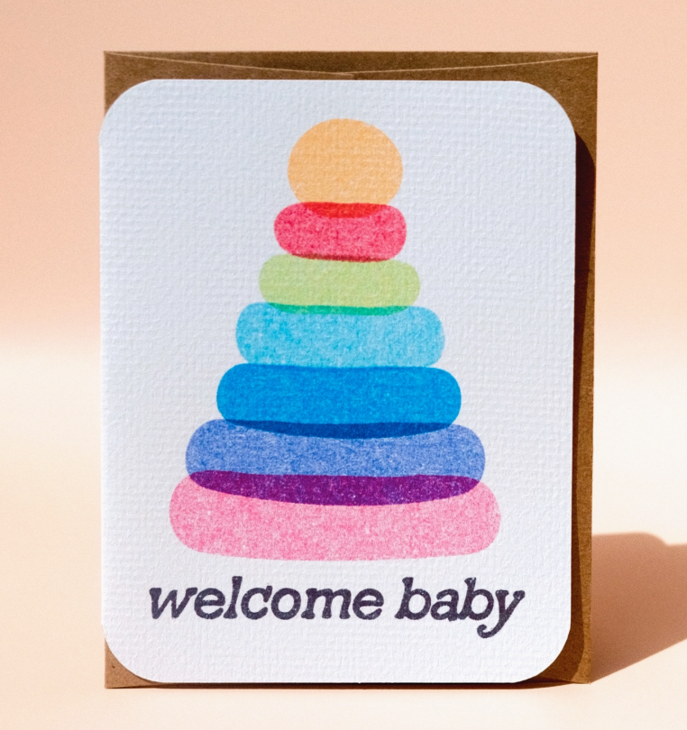 The Welcome Baby Card by Paper Chain Studios features a colorful stacked ring toy illustration with "welcome baby" on a textured white background, rounded corners, and comes with a brown envelope.