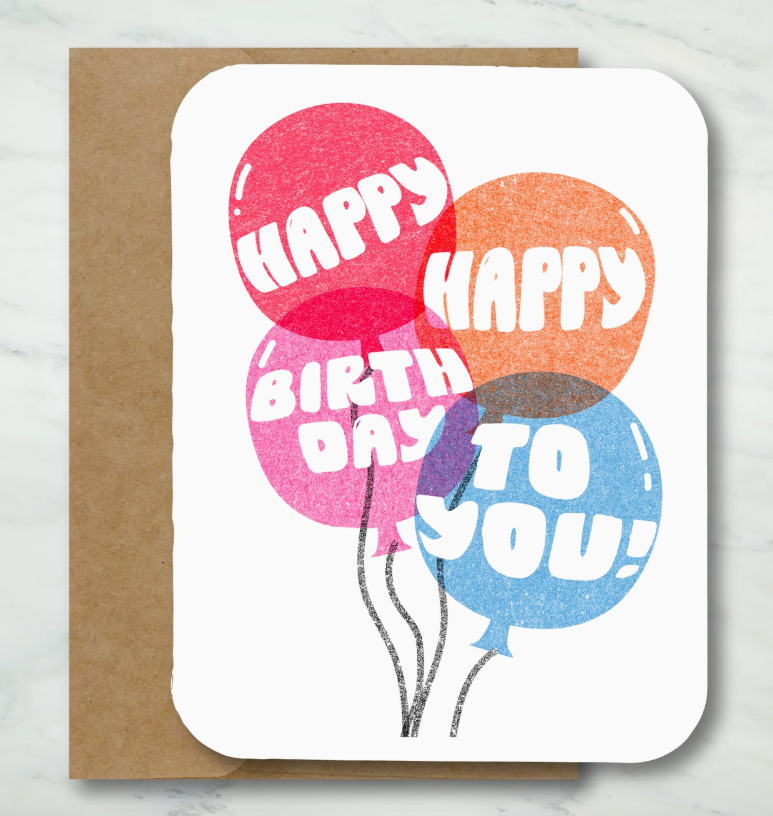 The Colorful Birthday Balloon Card by Paper Chain Studios features four vibrant balloons with “Happy Happy Birthday To You!” on the front, displayed atop a brown envelope against a marble background.