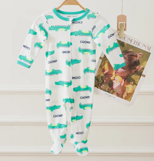A Lollipop Dreams Chompy Gator Zip Footie—white with green alligator prints and "CHOMP" text—hangs on a hanger beside a wall-clipped magazine featuring a woman eating fruit on its cover.
