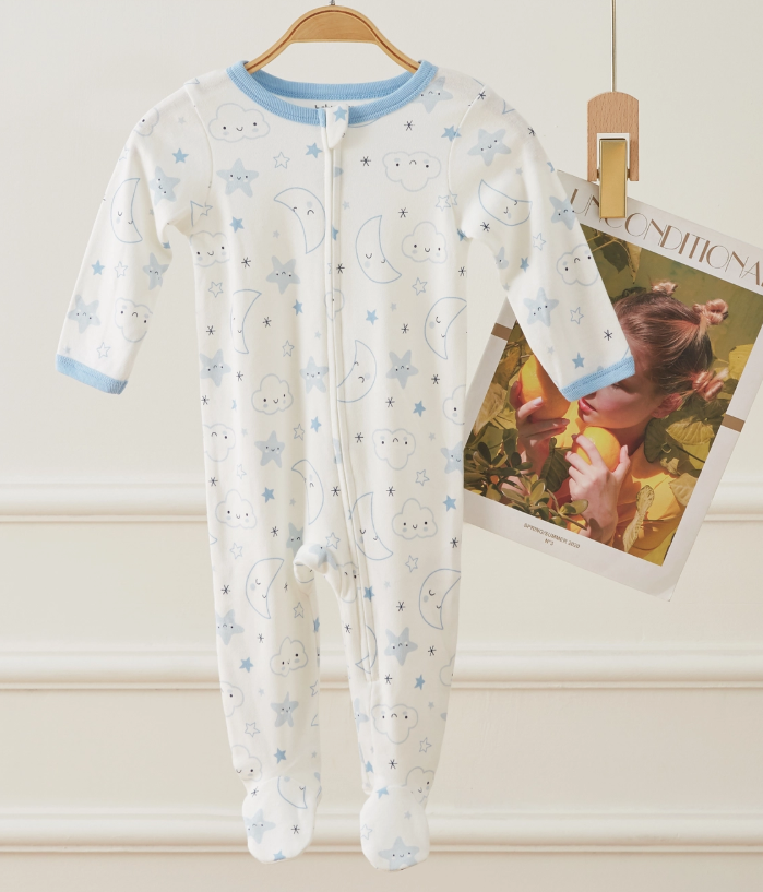 The Moonlit Dreams Zip Footie by Lollipop Dreams, a white onesie with blue trim and a celestial print, hangs on a wooden hanger next to a magazine page featuring a child with flowers.