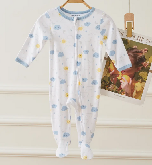 The Sunny Skies Zip Footie by Lollipop Dreams, a white baby onesie with blue trim, blue clouds, and yellow suns, hangs on a hanger beside a magazine featuring a woman holding fruit against a cream-colored wall.