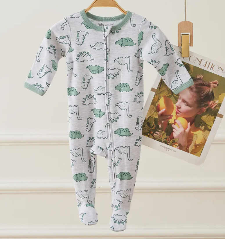The Lollipop Dreams Dino Doodles Zip Footie, a gray baby onesie with green trim and dinosaur print, hangs on a wooden hanger next to a magazine featuring a woman holding oranges.