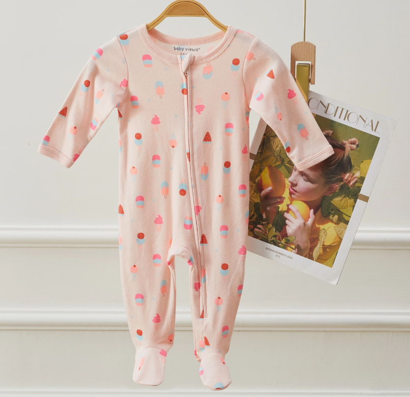 The Lollipop Dreams Sweet Treats Zip Footie, a pink onesie with colorful ice cream prints, hangs on a wooden hanger. Behind it, a magazine showing a woman with yellow fruit is clipped to the hanger against a white wall.