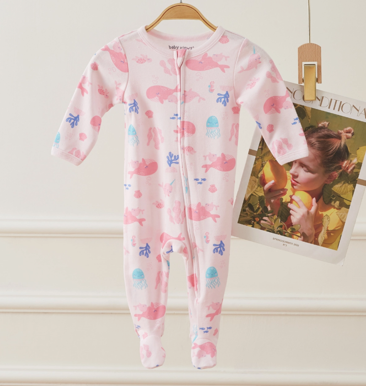 The Pink Sea Dreams Zip Footie by Lollipop Dreams, featuring blue and pink sea animal prints, hangs on a wooden hanger next to a magazine page of a child holding fruit near her face.