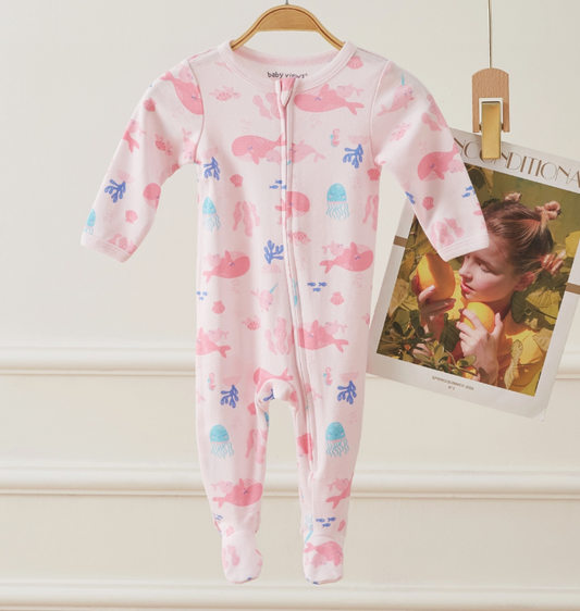 The Pink Sea Dreams Zip Footie by Lollipop Dreams, featuring blue and pink sea animal prints, hangs on a wooden hanger next to a magazine page of a child holding fruit near her face.