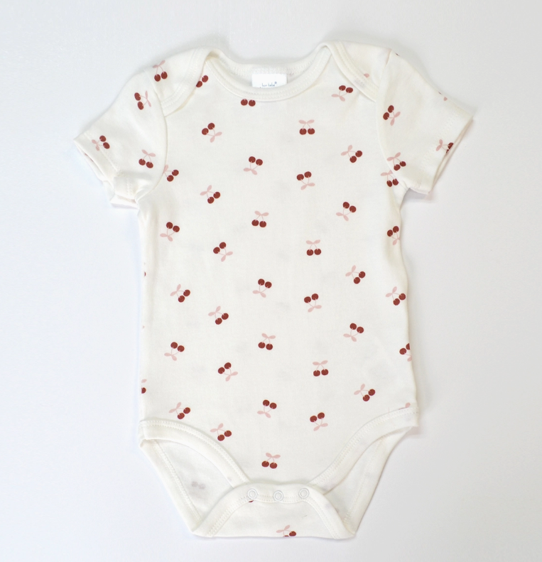 The Cherry Bow Bubble Trio by Lollipop Dreams is a white short-sleeve baby onesie featuring a repeating red cherry pattern and snap buttons at the bottom for easy changing.
