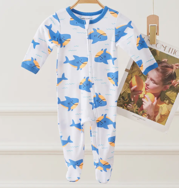 The Lollipop Dreams Shark Splash Zip Footie, a white baby onesie with blue trim and blue-orange cartoon sharks, hangs on a wooden hanger. A magazine page showing a woman eating a peach is in the background.