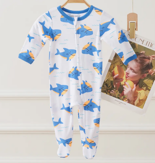 The Lollipop Dreams Shark Splash Zip Footie, a white baby onesie with blue trim and blue-orange cartoon sharks, hangs on a wooden hanger. A magazine page showing a woman eating a peach is in the background.