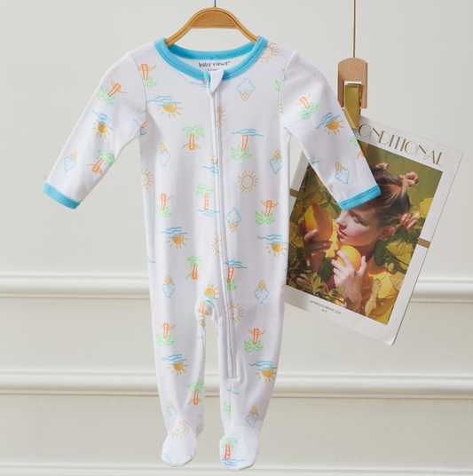 The Lollipop Dreams Sunny Days Zip Footie, a white baby onesie with blue trim and colorful suns, ice cream, and surfboards, hangs on a wooden hanger. Behind it is a partially visible magazine featuring a woman with a lemon.