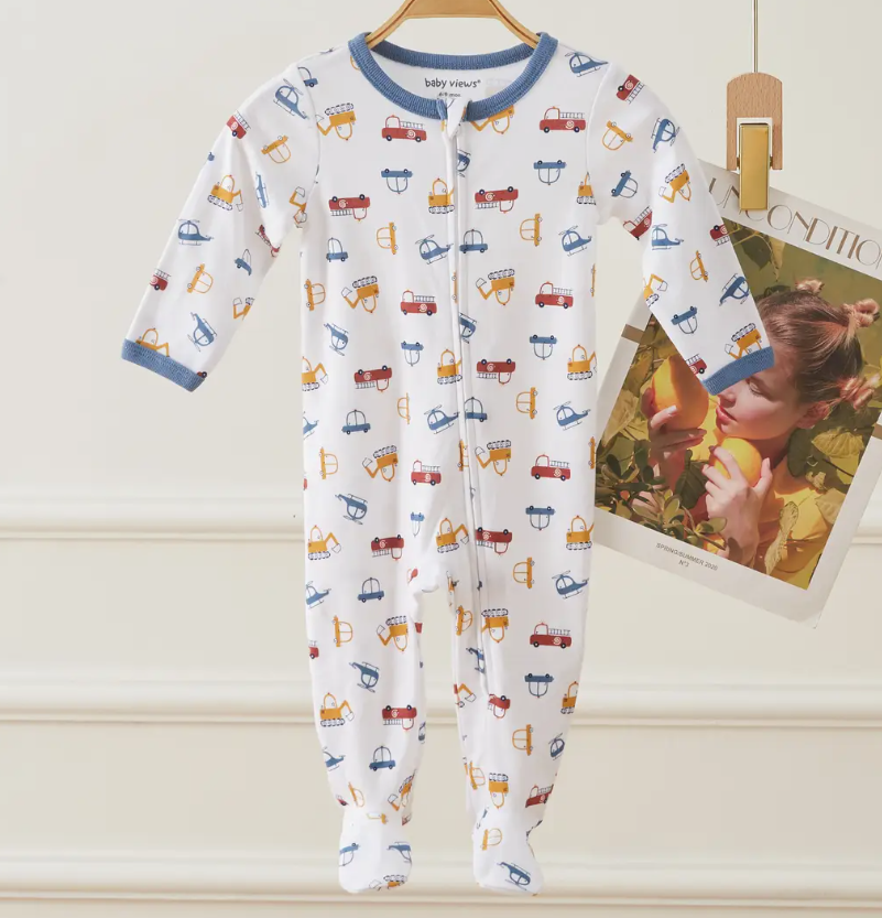 The Rescue Rides Zip Footie by Lollipop Dreams, a white baby onesie with blue trim and colorful construction vehicles, hangs on a wooden hanger beside a magazine showing a girl with an apple.