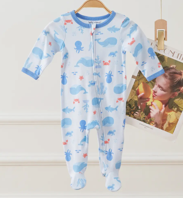 The Ocean Pals Zip Footie by Lollipop Dreams, featuring blue trim and an ocean animal print, hangs on a wooden hanger. Next to it, a magazine with a cover photo of a woman eating fruit is clipped for display.