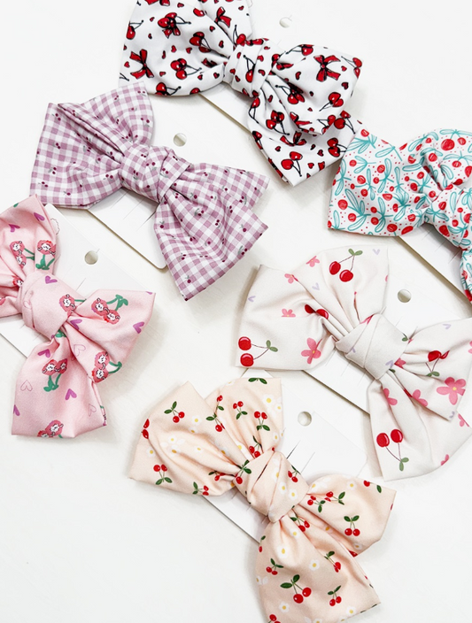The Sweet Cherry Days Hair Bow Clips by love & repeat feature six colorful fabric bows with cherries, flowers, and plaid patterns, displayed on white cards against a white background.
