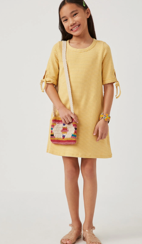 A smiling young girl poses against a white background wearing the Hayden LA Sunshine Stripe Dress—yellow and white stripes with short sleeves tied at the ends—paired with tan sandals, a colorful woven bag, and a hair clip.