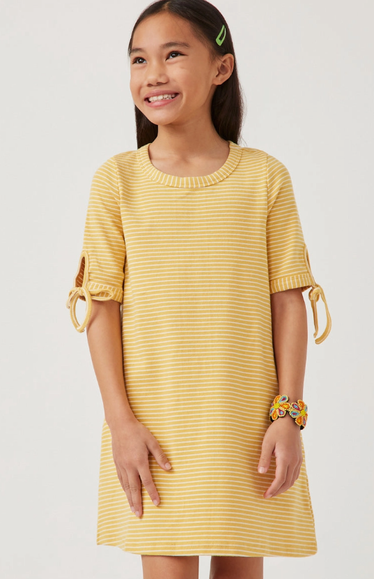 A young girl smiles in the Hayden LA Sunshine Stripe Dress, featuring yellow and white stripes and short sleeves with bows. She wears a green hair clip and a colorful butterfly bracelet, completing her cheerful look.