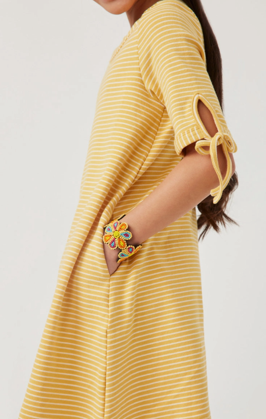 A girl wears the Hayden LA Sunshine Stripe Dress, a yellow and white striped short-sleeve dress with tie details and pockets, accessorized with a colorful beaded flower bracelet.