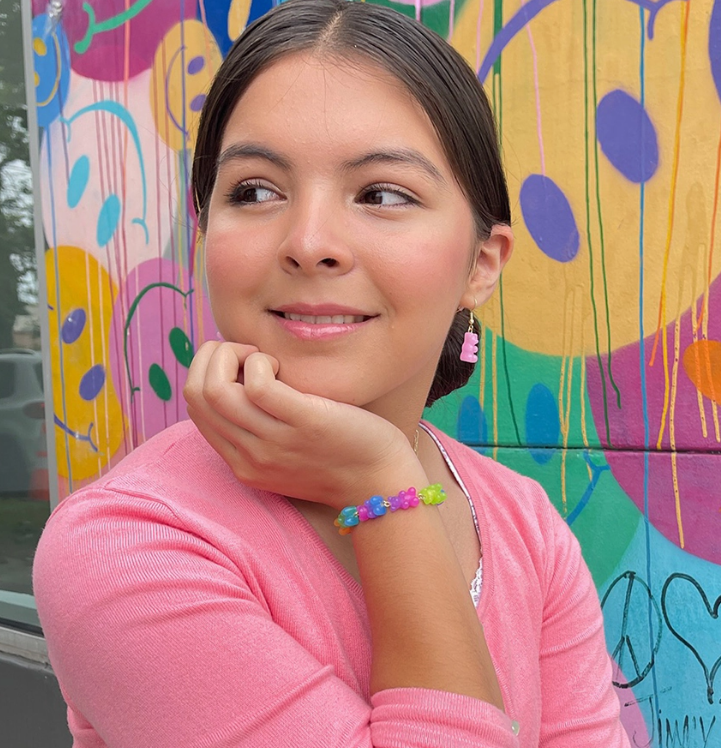 A young woman in a pink top, smiling gently with her chin on her hand, showcases the Lil Bear Thang Bracelet by top trenz inc. as she sits before a vibrant mural of abstract shapes and colors.