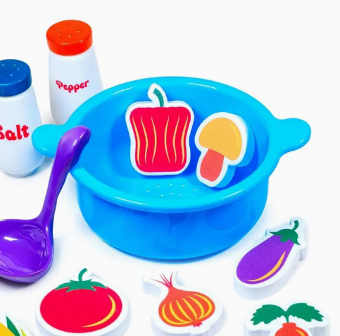 The TubWorks Tub Works® Tub Soup™ Scoop, Strain & Play Toddler Bath Toy features a blue bowl, purple ladle, wooden toy veggies—bell pepper, mushroom, onion, tomato, eggplant—plus salt and pepper shakers.