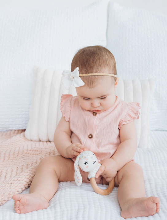 A baby in a pink outfit with a white bow headband sits on a bed, holding the PearHead Floral Snuggle Bunny Teether. White pillows and a pink blanket are visible in the background.