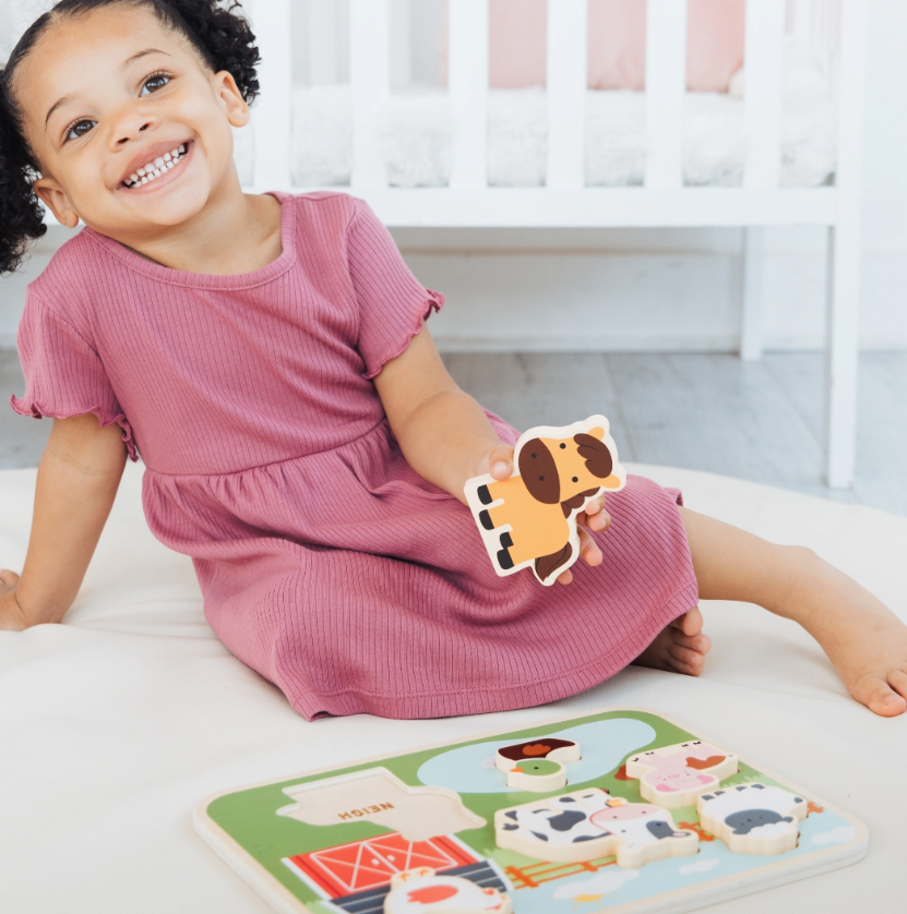 A smiling young girl in a mauve dress sits on the floor holding a piece from the PearHead Wooden Barn Animal Puzzle, with the puzzle board in front of her and a white crib in the background.