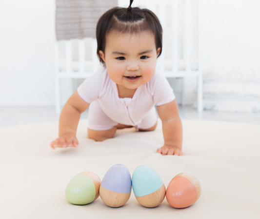 A baby in a light pink outfit crawls toward four pastel-colored PearHead Wooden Egg Shakers Toys, with a crib and soft blankets in the background.