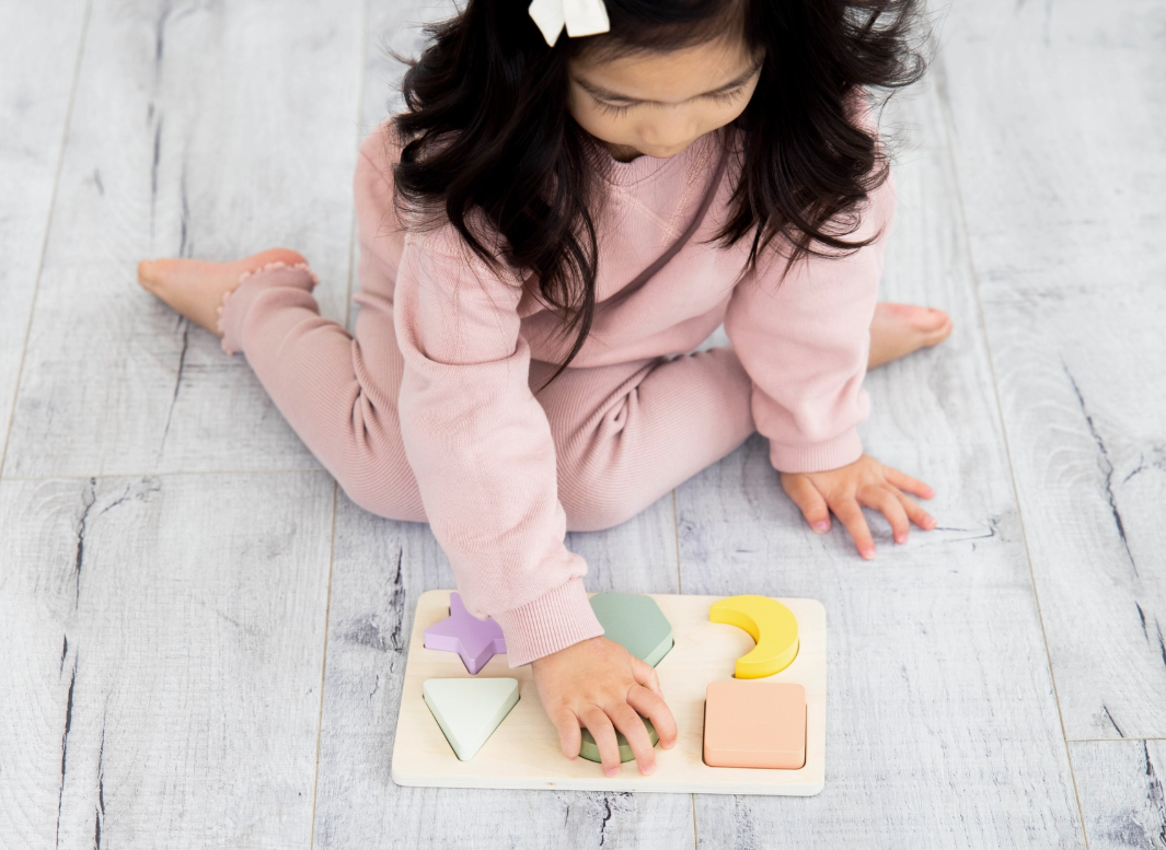 A young child in a pink outfit sits on a light wooden floor, playing with the PearHead Wooden Shapes Puzzle featuring pastel-colored geometric pieces.