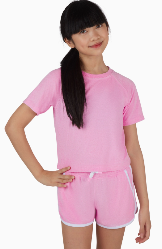 A young girl with long black hair in a ponytail smiles with one hand on her hip, wearing the 90 Degree by Reflex & Yogalicious Cotton Candy Towel Terry Short Set against a white background.
