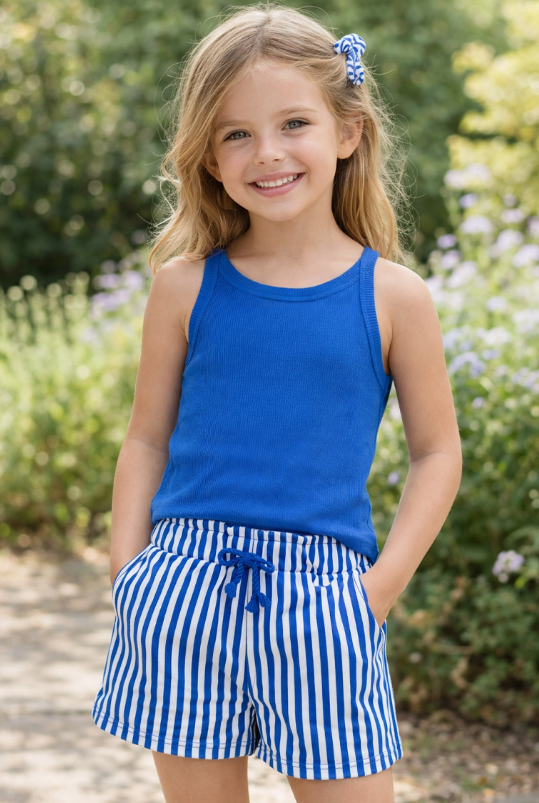 A young girl in the Cutie Patootie Blue Sporty Tank Set smiles outdoors with her hands in her pockets, surrounded by green foliage.