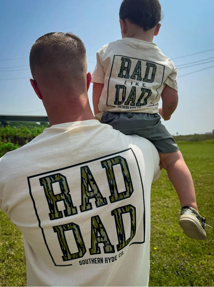 A man carries a child outdoors; both wear white Rad Dad Men T-Shirts by Wild Trail Outfitters with bold camo-style text—the man’s reads “RAD DAD,” the child’s says “RAD LIKE DAD.” They stand on green grass under a sunny sky.