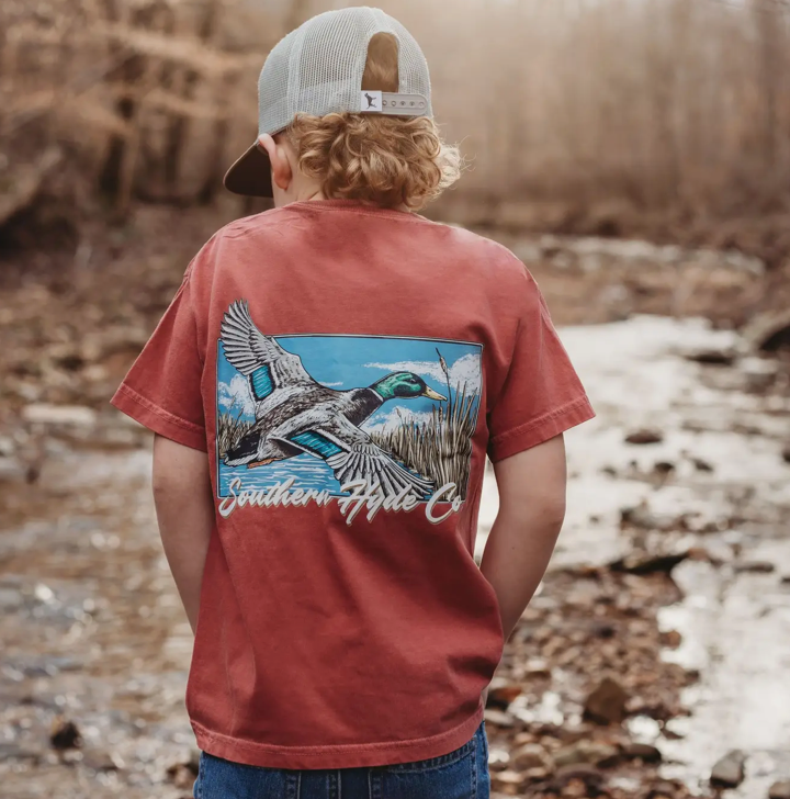 A child wearing the Wingspan Men T-Shirt by Wild Trail Outfitters—with a duck graphic—stands by a rocky creek in the woods, facing away from the camera and wearing a light-colored cap.