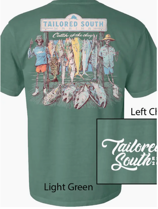 The Skeleton Fish Men T-Shirt by Wild Trail Outfitters features a light green color with a large back graphic of three people under a “Tailored South Marina” sign posing with big fish, and the “Tailored South” logo in white on the front left chest.