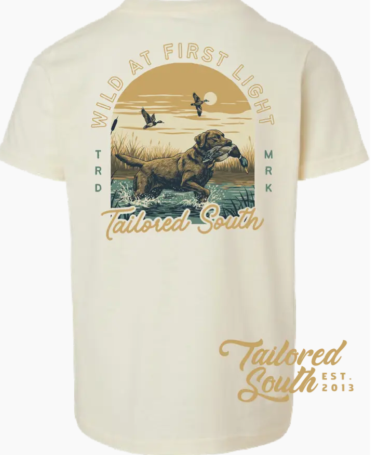 A cream T-shirt featuring a dog retrieving a duck, birds flying, and sunrise art. Text: WILD AT FIRST LIGHT, Wild Trail Outfitters, and EST. 2013. Youth size; style: Wild At First Light Youth.