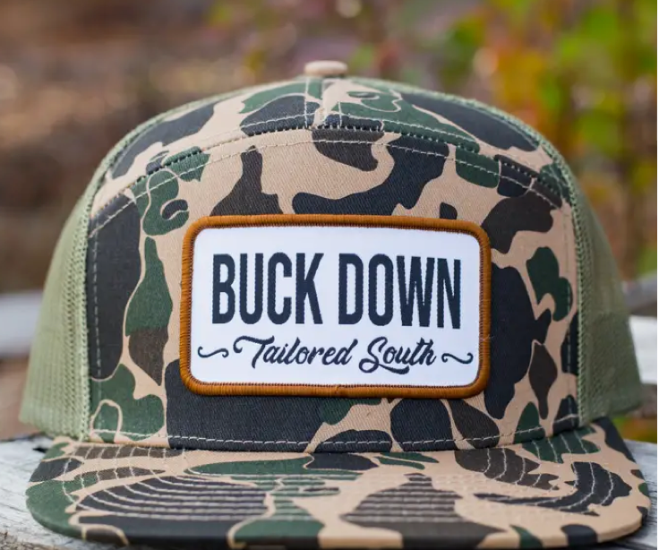 The Buck Down Adult Hat by Wild Trail Outfitters features a camouflage trucker design with a front patch that says BUCK DOWN in bold and Tailored South in script underneath.