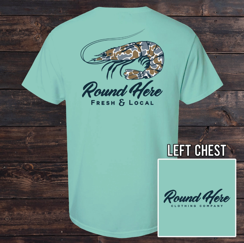 The Old North State Fresh & Local Shrimp Men's Tee features a light blue camo-patterned shrimp graphic and “Round Here Fresh & Local” on the back, with the brand logo on the front chest. Displayed on a wooden background.
