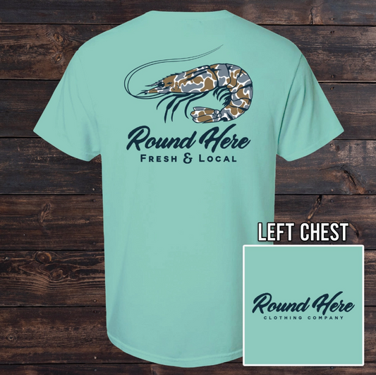 The Old North State Fresh & Local Shrimp Men's Tee features a light blue camo-patterned shrimp graphic and “Round Here Fresh & Local” on the back, with the brand logo on the front chest. Displayed on a wooden background.