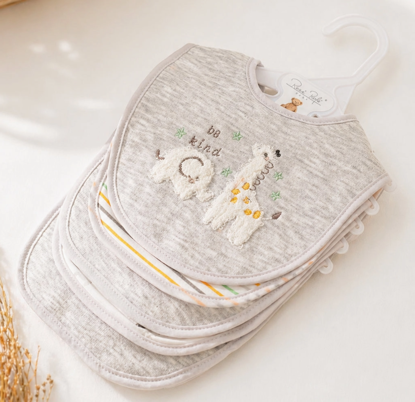 A stack of Lollipop Dreams Be Kind Llama Bib Set in gray, featuring embroidered animals and "be kind" on the front, hangs on a white plastic hanger, displayed on a light surface.