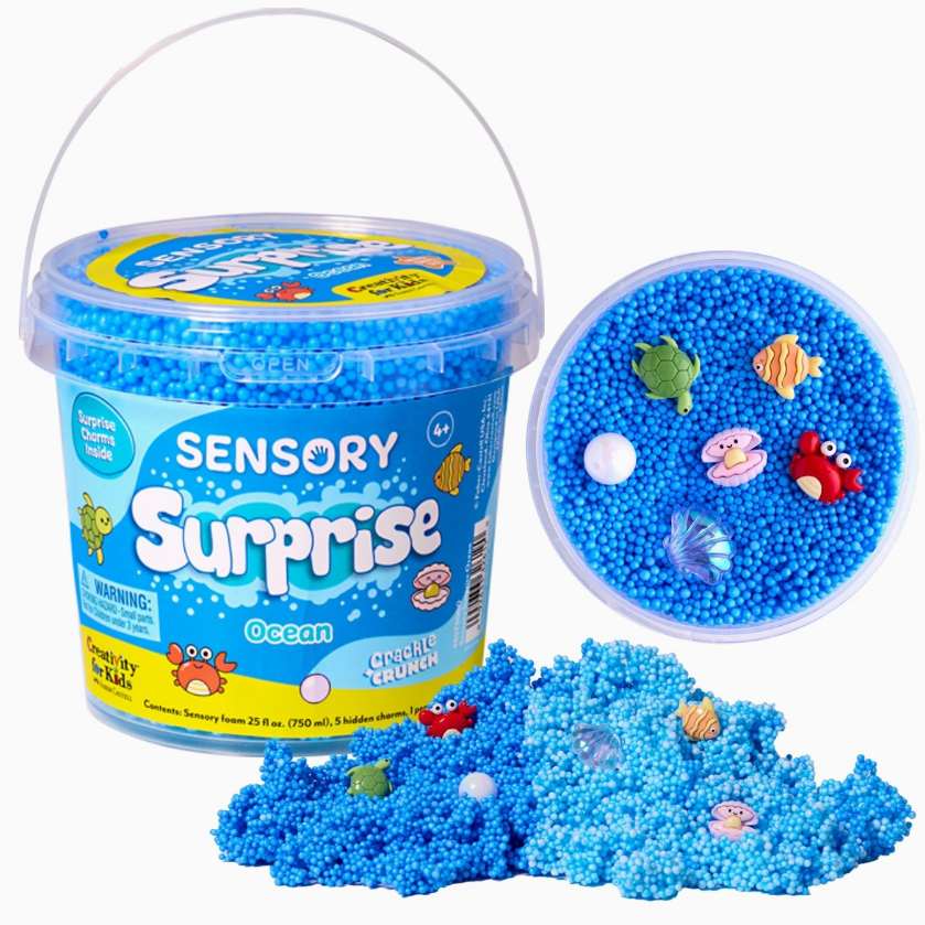 The Creativity For Kids Sensory Surprise Squishy Foam Bucket includes blue foam beads, ocean-themed charms, and a matching lid. Beads and charms are pictured both spilled out and inside a separate round container.