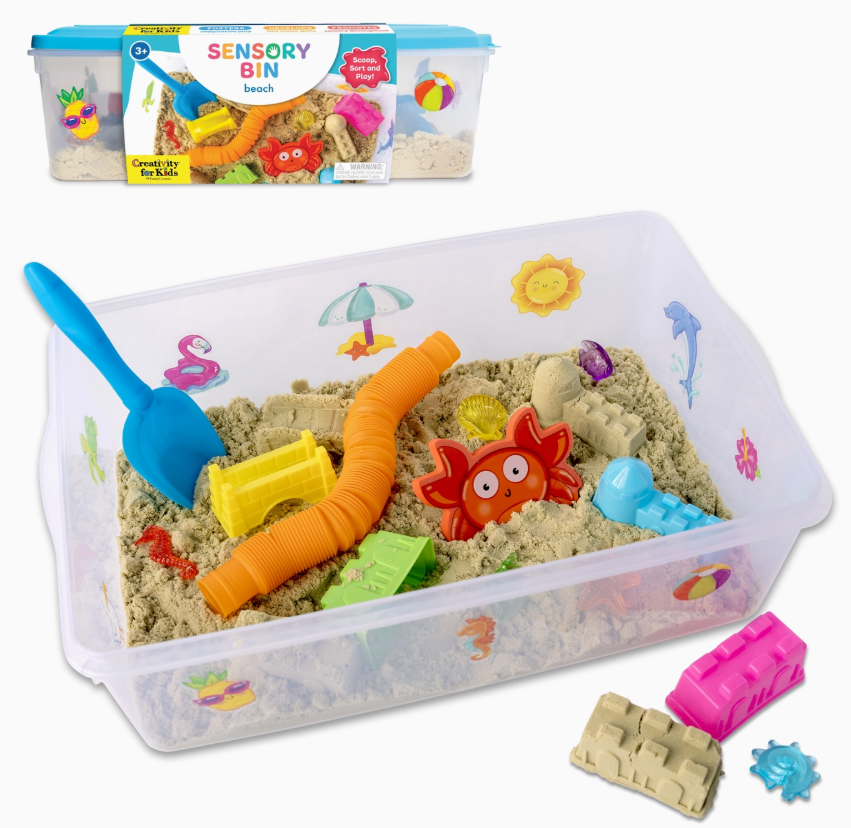 The Beach Sensory Bin by Creativity For Kids includes sand, a blue shovel, an orange tube, red crab, yellow and green molds in a clear bin decorated with sea and sun stickers. The product box is visible in the background.
