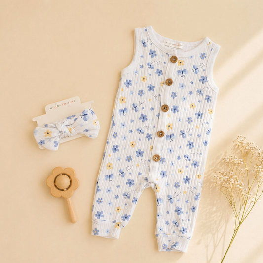 The Little Blue Meadow Set by Lollipop Dreams features a sleeveless baby romper with blue and yellow floral print, front brown buttons, a matching headband, wooden flower-shaped rattle, and dried flowers on a beige background.