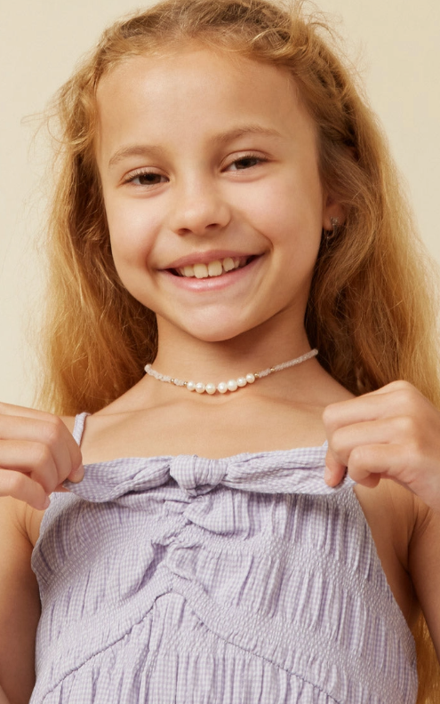 Smiling young girl with long blonde hair wearing the Hayden LA Purple Bow Tank Romper and a beaded pearl choker necklace, holding the romper's straps with both hands.